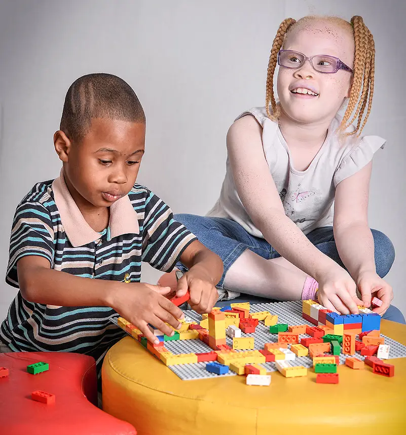 Foto de duas crianças, um menino e uma menina, brincando com peças coloridas do LEGO Brille Bricks em cima de uma bancada amarela.